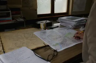 A city map printout, archival plan, and cup of tea on a municipal desk with a hand tracing a route, bathed in soft daylight.