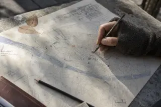 Close-up of a hand-drawn city map on a stone bench in a quiet urban square, a hand adds a colorful symbol, with markers, leaves, and pencil nearby in soft winter light.
