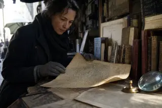 Antique market stall with a person studying old Paris maps in soft dawn light, detailed close-up on worn paper and objects.
