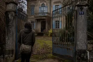 Researcher observes details of an old wrought iron gate and mossy threshold at the entrance of a Belle Époque villa in subdued winter light