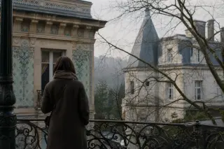 Contemplative person viewed from behind at ornate balcony, with blurred details of historic villas through bare branches in soft morning light.