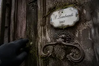 Close-up of a mossy old wooden door with a wrought iron handle, ornate ceramic nameplate, and a gloved hand reaching toward it in soft natural light.