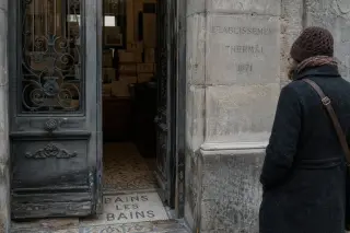 Weathered Belle Époque doorway with mosaic tiles, cast-iron grille, and blurred archive boxes inside; a lone person reads an inscription in diffuse winter light.
