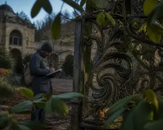 Oblique close-up of decorative iron gate with moss and blurred rhododendron leaves in foreground, solitary figure sketching architecture in soft morning light, contemplative and subtly nostalgic ambiance.