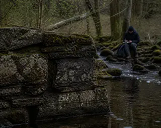 Mossy stone wall fragment in a forest stream, with a solitary person in the background writing in a journal, under soft overcast light.