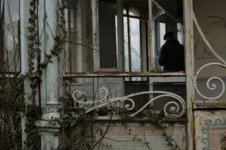 Detail of weathered ornate ironwork and faded mosaics on an abandoned villa veranda, ivy creeping in, with a solitary observer silhouetted through cracked glass, in muted winter light.