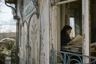 Fragment of a villa façade with art nouveau details and peeling paint, a woman inside studying archival documents, soft natural winter light, overgrown garden outside.
