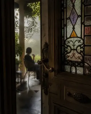 Subtle view of an old villa entrance with stained glass, a half-open door, and a solitary figure on a distant veranda in soft morning light