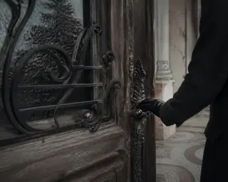 Gloved hand touches an ornate, weathered villa door with ironwork, soft winter light reflecting nearby trees, evoking quiet contemplation