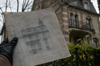 Close-up of a gloved hand examining an old architectural plan in front of a faded Belle Époque villa, with winter branches and ornate ironwork visible in soft daylight.