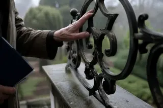 Close-up of an elderly hand touching ornate ironwork on a villa balcony, with dew and garden softly blurred in the background under grey, gentle morning light.
