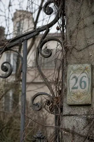 Detail of an Art Nouveau wrought-iron gate and ceramic street number, partly covered by winter vines, with a blurred villa façade in the background under soft overcast light.