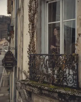 Faded villa balcony with wrought-iron details, moss, and pensive woman behind vintage glass on a quiet winter street, soft natural light