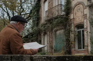 Detail of a weathered villa façade with Art Nouveau elements, partly covered by ivy, as an elderly resident reads an old map on a garden wall in soft morning light.