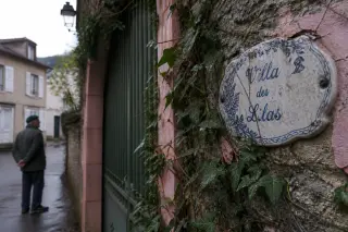 Weathered villa name plaque on stone facade partially covered by ivy, with an older local gazing quietly nearby, gentle morning light, subdued and contemplative mood
