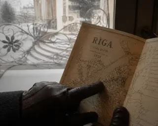 A hand in gloves examines an old map near a villa's decorated step, with a blurred ornate fence and subdued winter light.
