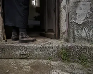Weathered chapel threshold with moss, wildflowers, and the feet of an elderly person pausing in soft grey morning light