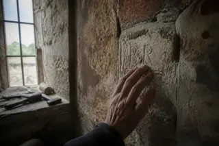 Detail of an old stone church wall with faded ochre, an artisan's hand tracing engraved initials, and old masonry tools on a ledge in soft grey daylight.