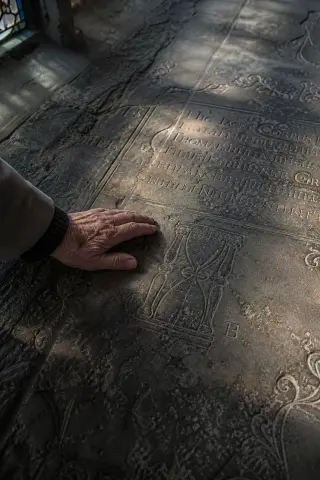 A close-up of old stone plaques with faint inscriptions on a church floor, touched gently by a person's hand in soft, wintry light.