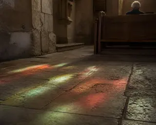 Colored reflections from stained glass illuminate old church floor, with a solitary figure seated pensively in the background.