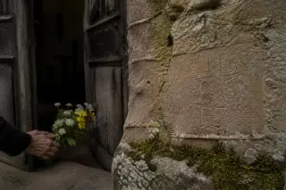 Close-up of old sandstone chapel wall with moss and faded inscription, while blurred hands leave wildflowers on the threshold in soft, natural light