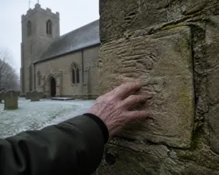 Elderly hand tracing chiseled stone block on old rural church wall, in soft winter light, with moss and subtle historic texture