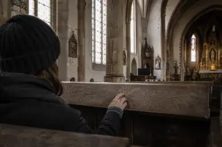 Person contemplatively touching carved initials on a wooden church pew, subtle stained glass light and gold details in background, soft natural lighting.