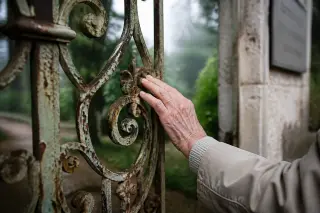 Hand of elderly person touching weathered Belle-Époque iron gate with soft light and blurred historic park background