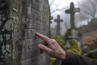Weathered hand gently tracing names on a mossy headstone in a quiet, wintery cemetery, soft light and blurred gravestones in the background.
