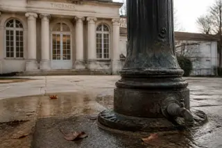 Worn iron pump base on damp stone pavement, scattered autumn leaves, ornate neoclassical façade and large arched windows of thermal baths in the soft winter light.