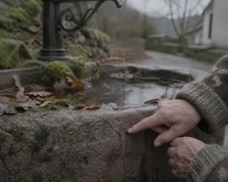 A close-up of an elderly person's hand touching the engraved date on a mossy, rain-soaked stone basin in a rural village, with fallen leaves and a blurred fountain spout in the background under soft, grey winter light.