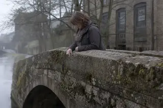 Woman absorbed in examining time-worn inscription on mossy canal bridge, industrial building and trees blurred in misty spring light