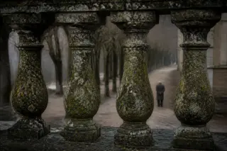 Detail of a moss-covered old stone balustrade overlooking a blurred, tranquil park alley with a solitary, contemplative figure in the distance