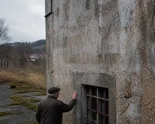 A person in a coat touches an old, weathered factory wall with faded lettering and moss, softly lit on a wintry day, behind them an overgrown industrial lot.