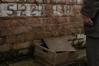 Fragment of an old brick wall with moss and faded numbers, a wooden archive box on the ground, and the shadow of a solitary person nearby in soft winter light.