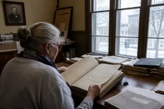 An elderly woman studies old documents in a softly lit archive room, surrounded by historic papers and a faded portrait, with cold daylight entering through antique windows.