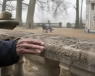 A hand touches the weathered, mossy balustrade of a historic spa park, with delicate architectural details and a solitary reader on a bench in the background among bare trees.