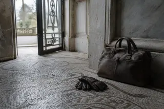 Worn mosaic floor with a leather bag and gloves beside an open Art Deco door, hinting at a park beyond, in soft winter daylight.
