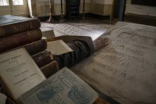 Elderly woman's hand tracing engraved stone plaque in a dim archive, surrounded by old registers and documents, soft winter daylight highlighting the scene.