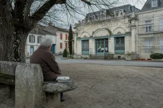 Elderly woman in winter coat sits alone on a stone bench by a Belle Époque spa building with weathered details and newsletters, under overcast soft light in a quiet village square.