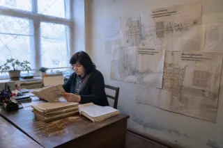 Municipal records room with a woman examining files, faded maps of villages on the wall, winter daylight and a quiet, introspective atmosphere.