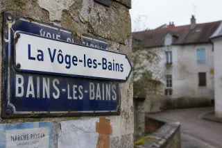 Partial street sign overlay with both new and old village names visible, set against a damp stone wall and community notice fragments under soft winter light.