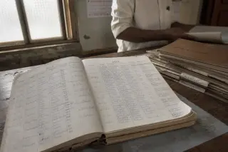 Handwritten old register book open on wooden desk, soft daylight, and a person in the background sorting labeled folders, evoking quiet administrative history.