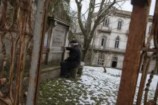 Elderly person in winter clothing sitting alone on a stone bench, tracing names on a weathered plaque in an overgrown courtyard of a faded thermal building, soft grey daylight, subtle architectural details, framed through a rusty iron gate.