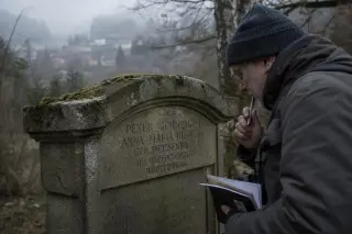 Researcher examining an old gravestone with foreign surnames in a misty Vosgian cemetery near a blurred forest edge