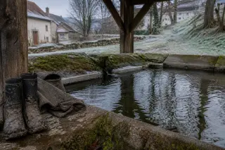 Weathered rural washhouse basin with mossy stones, muddy boots and a wool jacket near the water, under soft grey winter light.