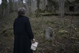 A woman in a dark coat examines a mossy memorial stone near the edge of a quiet Vosges forest in winter, holding an old letter.