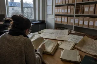 A person absorbed in studying medieval manuscripts at an archive table, with soft daylight highlighting the texture of ancient documents and a tranquil, introspective mood.