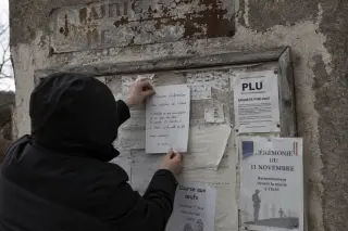 Detail of an old notice board with overlapping faded flyers and plaques, while a solitary person adds a handwritten note, all under soft grey winter light.