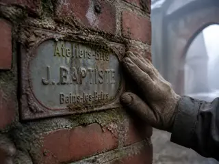 Old metal nameplate on a mossy, decaying brick wall, gently touched by a gloved hand, misty winter light hinting at abandoned industrial heritage